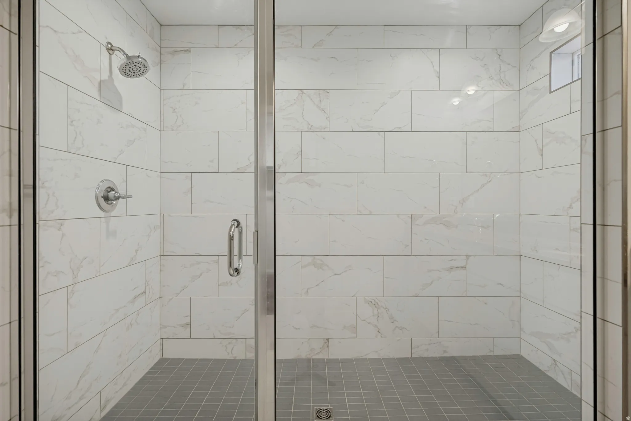 Glass-enclosed shower with white tiled walls, gray tiled flooring, and a chrome showerhead