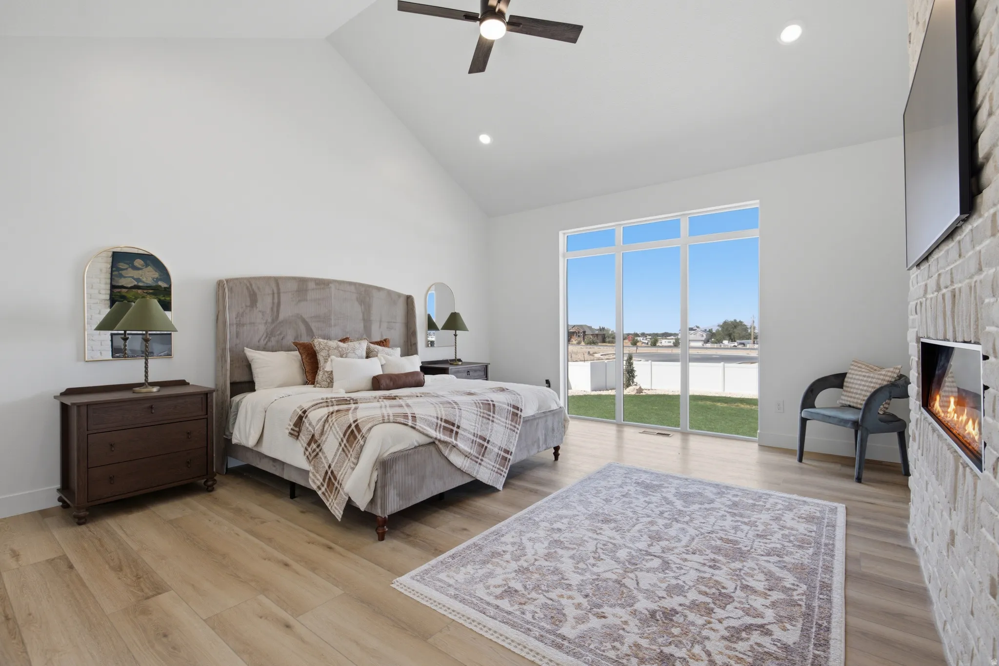 Vaulted ceilings, wood-finish flooring, a contemporary fireplace with a light brick surround, a ceiling fan, and expansive windows