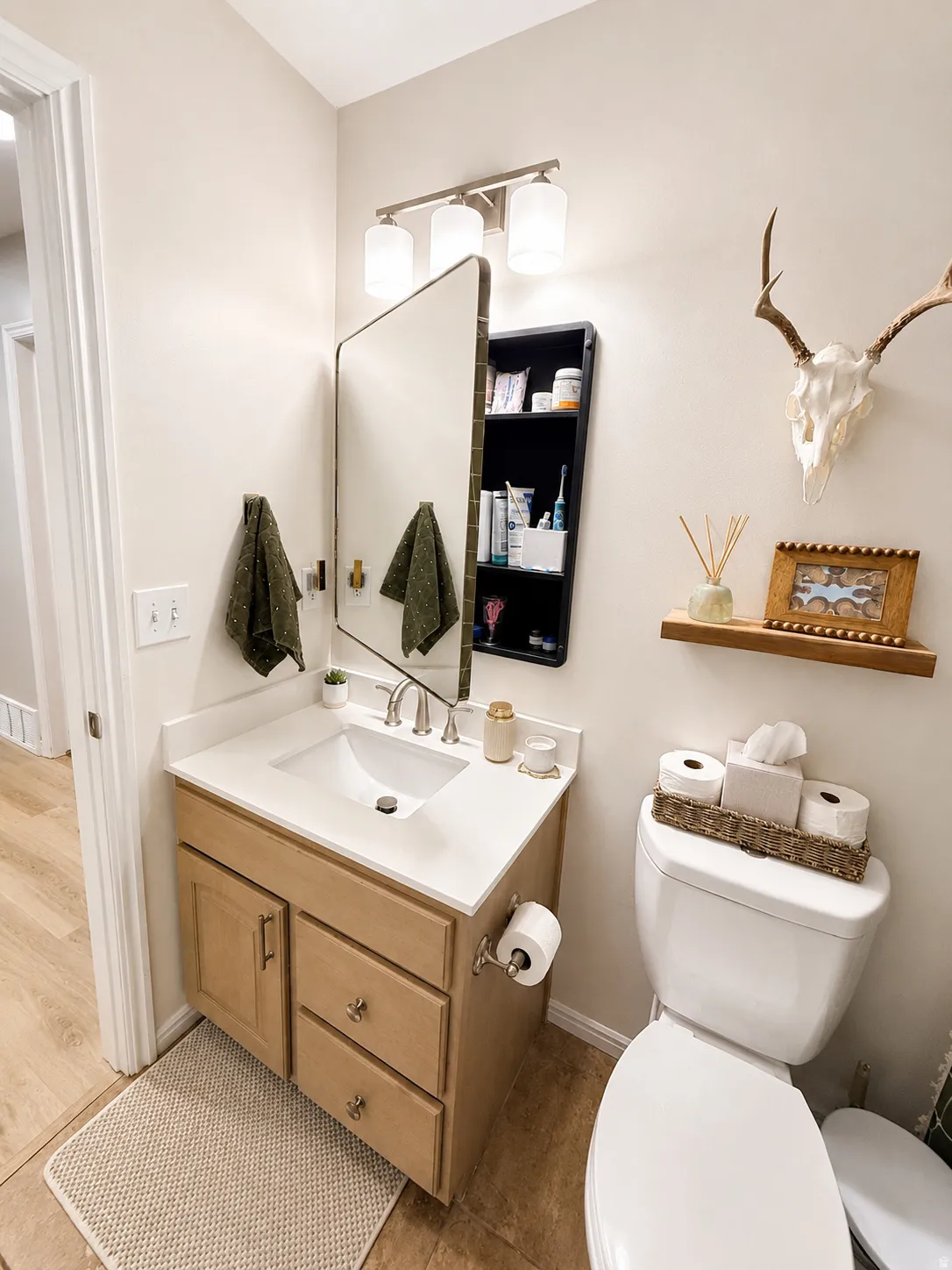 Single vanity with a light wood-finish cabinet and white countertop, undermount sink, three-light vanity fixture, and a wall-mounted mirror