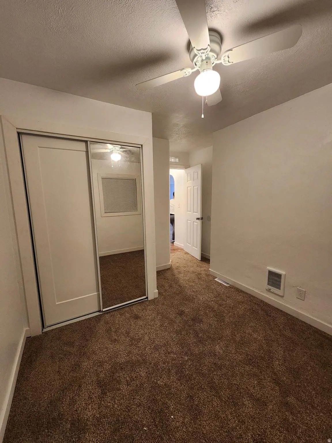 Sliding closet doors with mirrored panel, ceiling fan with integrated lighting, wall-mounted heating unit, and brown carpeting