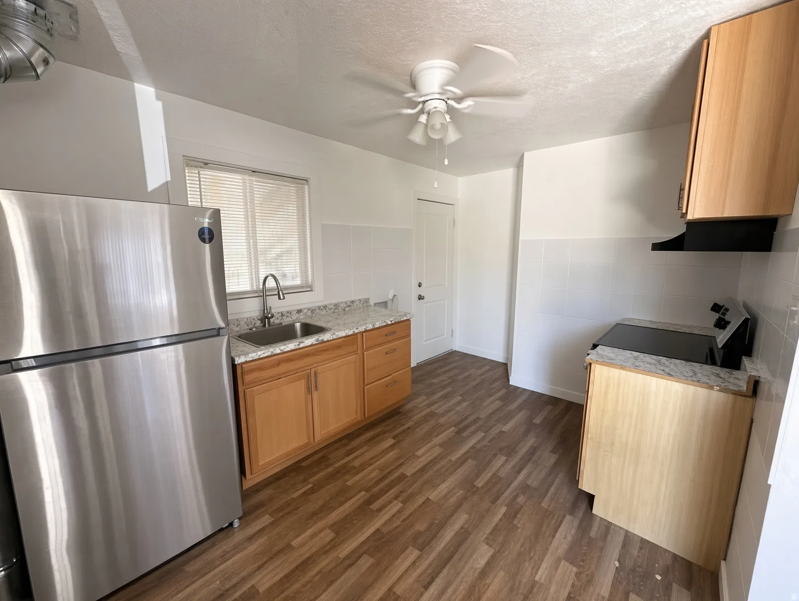 Stainless steel refrigerator, wood-finish flooring, light wood cabinetry, white tile backsplash, and a ceiling fan with light fixture