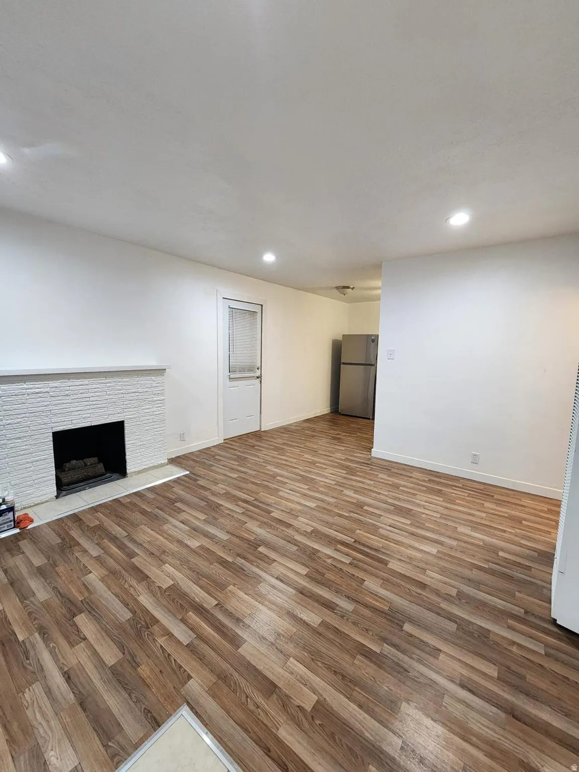 Wood-finish flooring, white brick fireplace with hearth, recessed lighting, and stainless-steel refrigerator