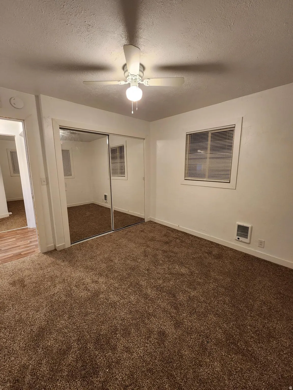 Neutral wall paint, ceiling fan with light fixture, mirrored closet doors, window with horizontal blinds, and wall-mounted heating unit