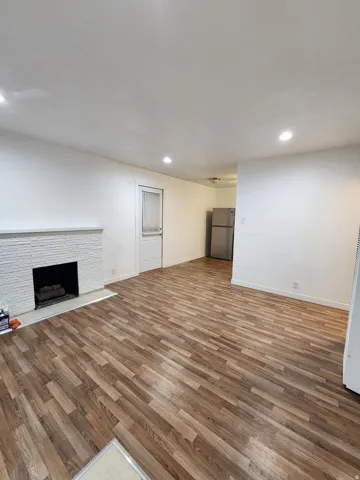 Wood-finish flooring, white brick fireplace with hearth, recessed lighting, and stainless-steel refrigerator