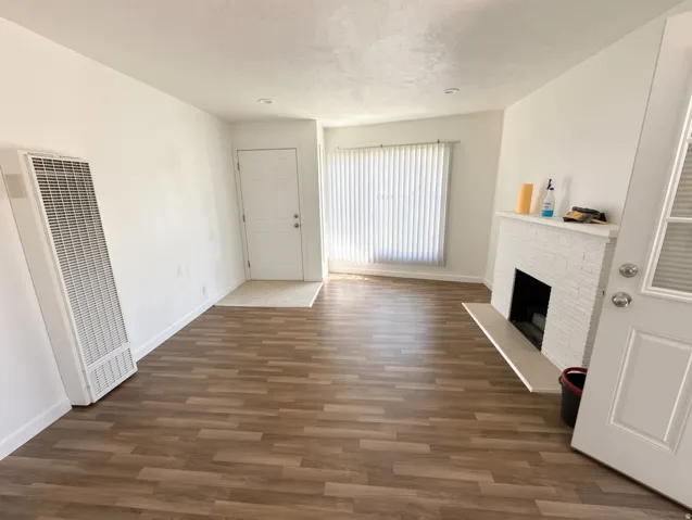 Wood-finish flooring, white painted brick fireplace with hearth, white window blinds, recessed lighting, and a wall-mounted heating unit