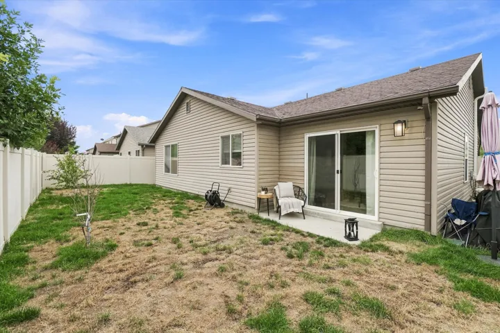 Spacious backyard with mature landscaping and established lawn area, complemented by a single-story residence featuring vinyl siding, multiple windows with dark frames, and a sliding glass door patio entrance