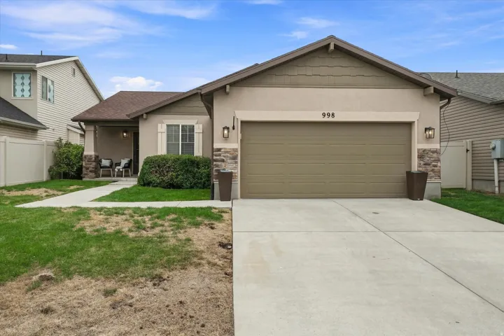 Charming single-story home featuring a two-car attached garage with a concrete driveway, neutral-toned exterior siding, stone accents flanking the entry, and manicured landscaping with established shrubs and lawn