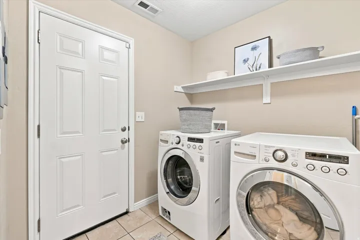Dedicated laundry room featuring two front-load washers with digital controls, tile flooring, floating shelving with decorative storage, and direct access via a six-panel entry door