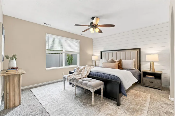 Spacious primary bedroom showcasing a black metal-frame bed with upholstered headboard, dual nightstands with integrated lighting, ceiling fan with light fixture, and large window with plantation shutters