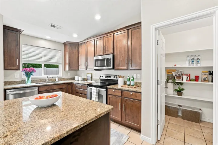 Chef's kitchen featuring warm wood cabinetry, stainless steel appliances, granite countertops, a center island with seating, and a convenient walk-in pantry with shelving and organizational storage