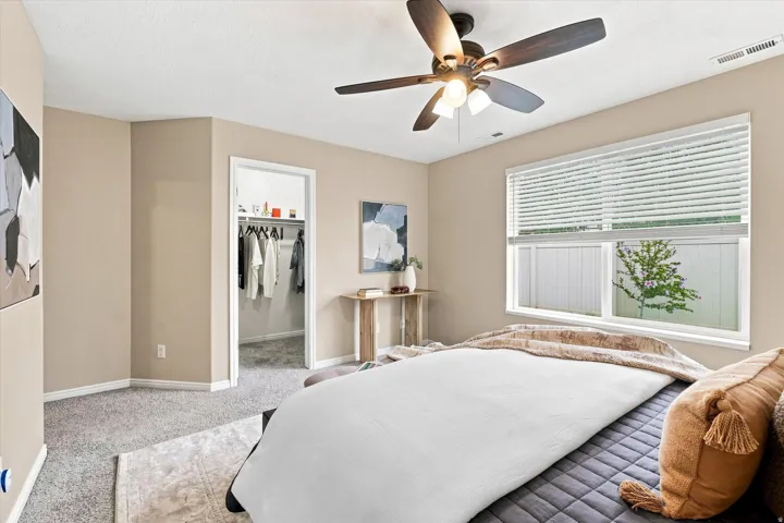 Master bedroom featuring neutral wall finishes, carpeted flooring, ceiling fan with integrated lighting, large windows with horizontal blinds providing natural light, and a walk-in closet with mirrored doors