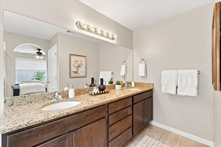 Spa-inspired primary bathroom featuring dual vanities with warm wood cabinetry, granite countertops, and polished brass hardware