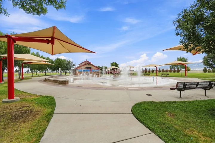 Community splash pad with multiple water features, shade structures, paved walkways, and integrated park benches