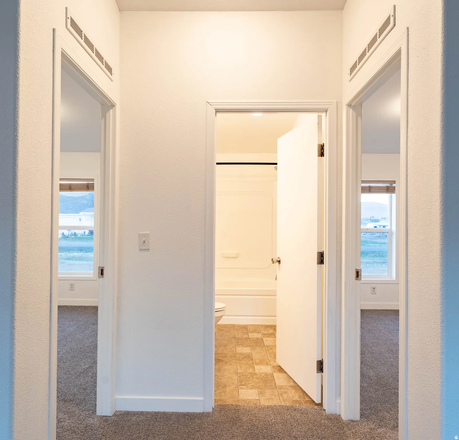 Hallway with neutral carpeting, leading to a bathroom with tile flooring and a bathtub/shower combination, and two rooms with neutral carpeting and windows