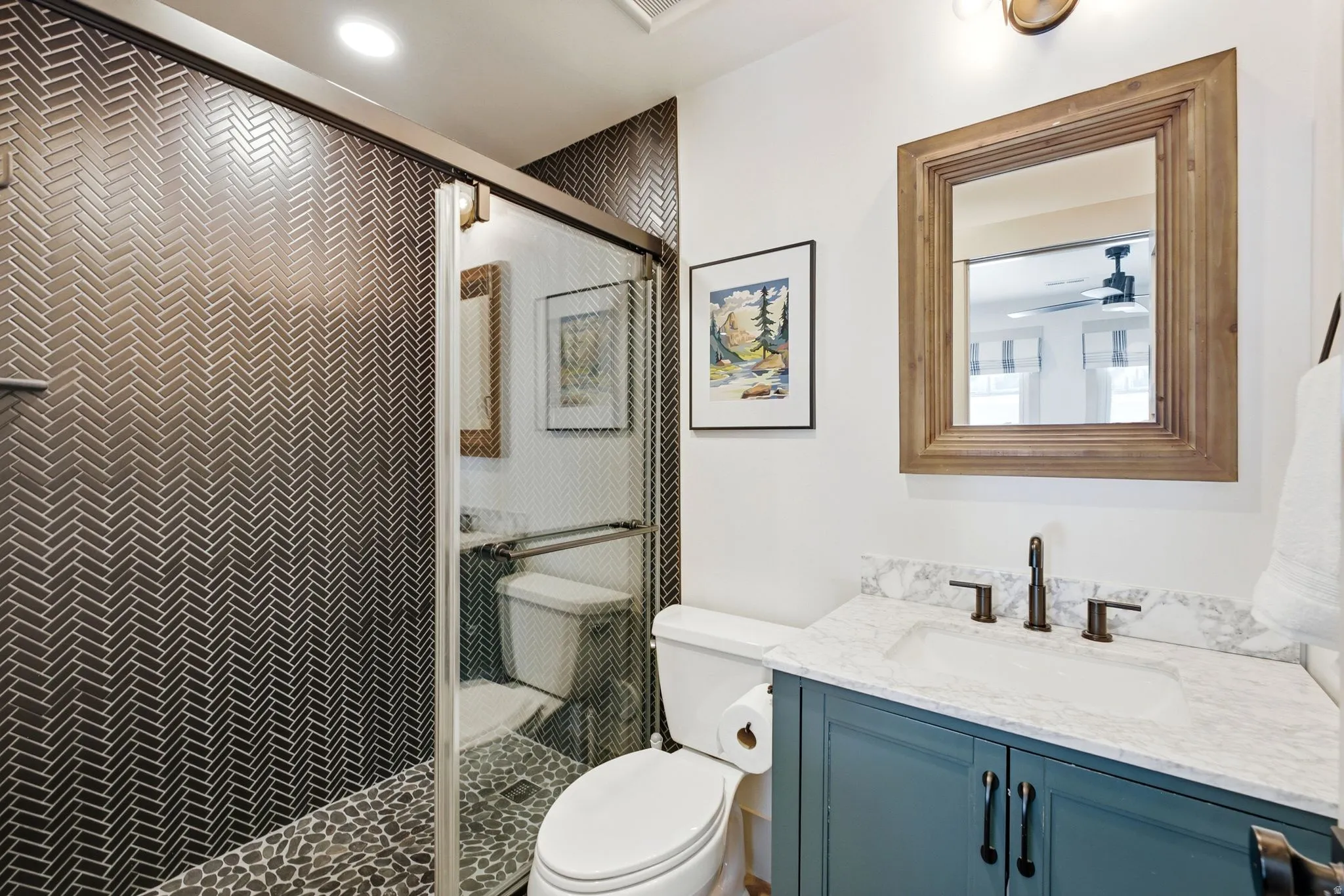 Herringbone-patterned shower tiling, pebble shower floor, glass shower enclosure, wood-framed mirror, and a vanity with a stone-finish countertop