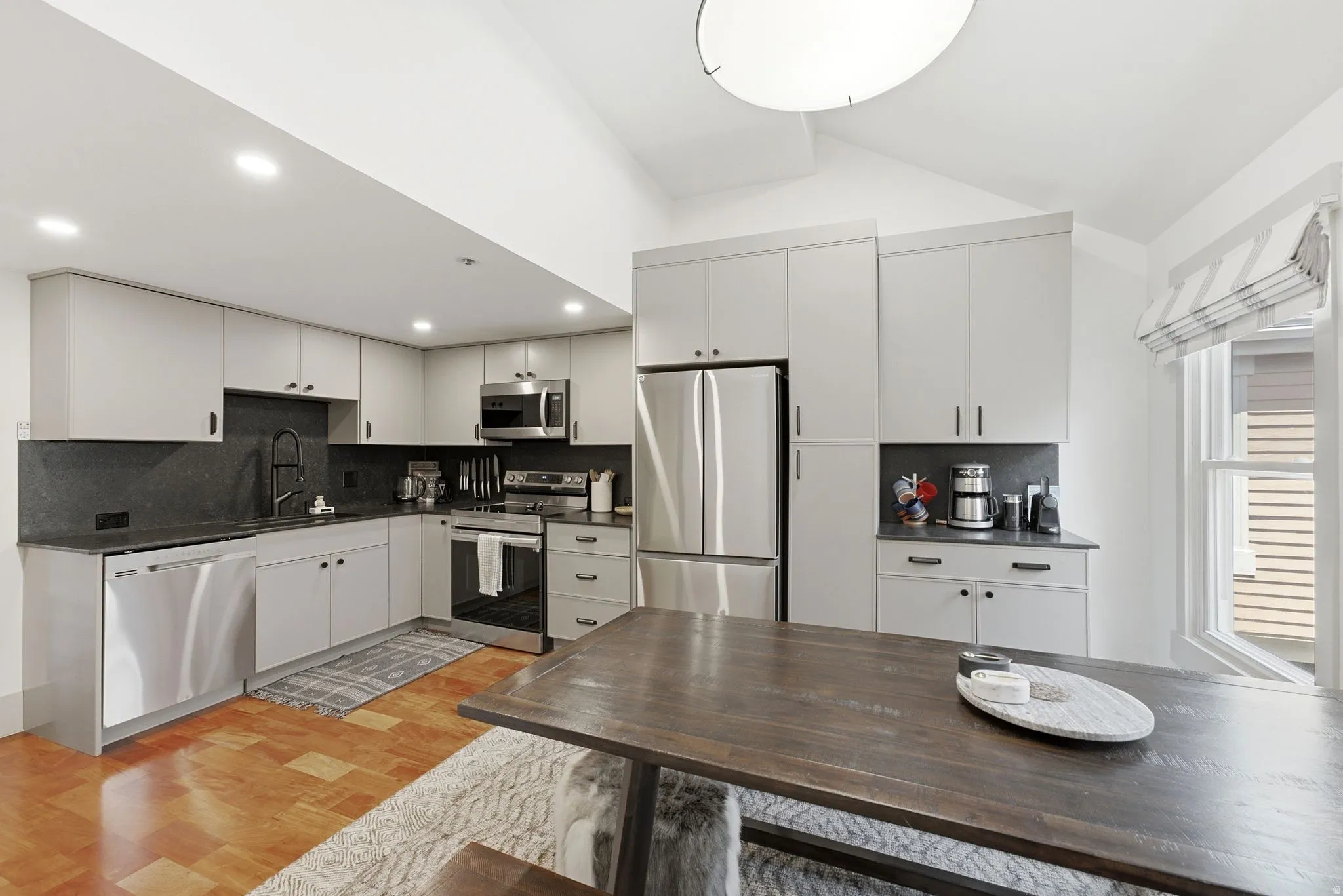 Modern kitchen featuring light gray cabinetry, stainless steel appliances, dark countertops with a coordinating backsplash, and wood-finish flooring