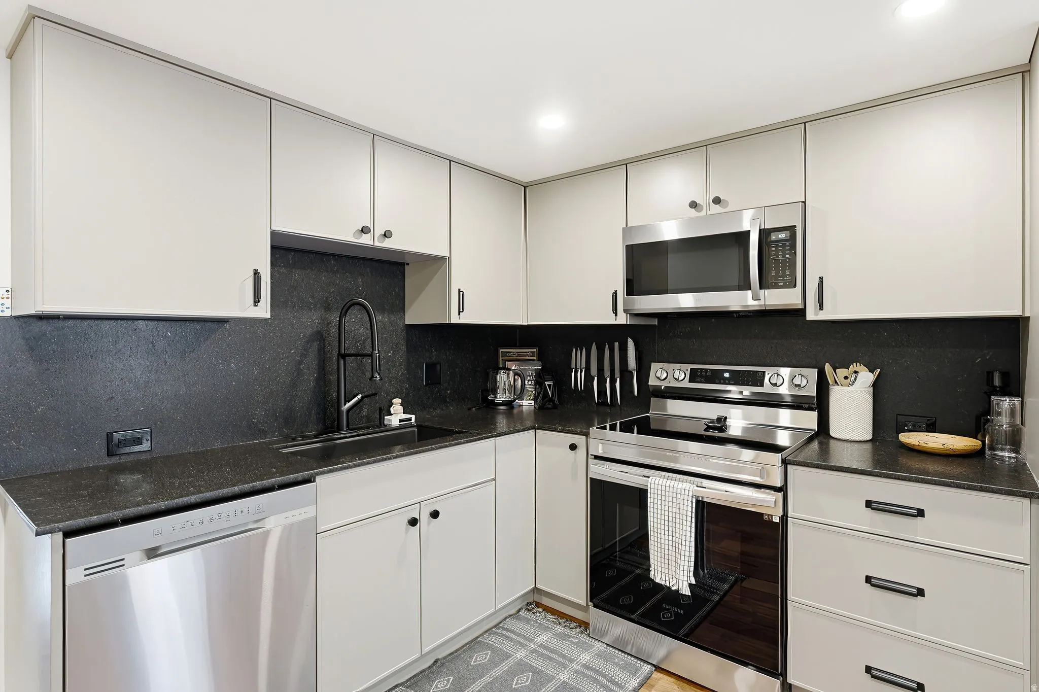 Contemporary kitchen featuring light gray cabinetry, dark stone countertops, stainless steel appliances, and a matte black faucet