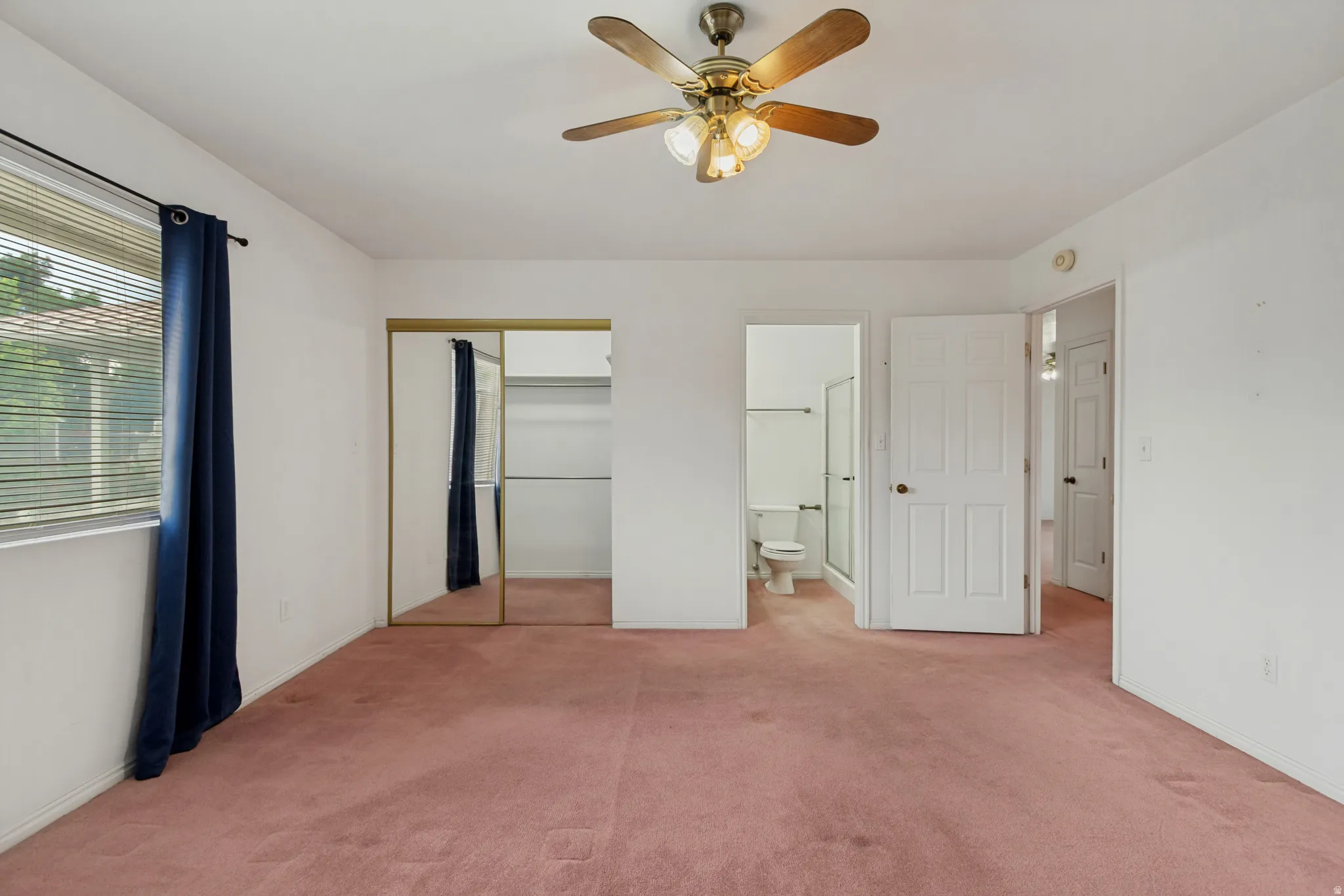 Primary bedroom shown vacant. Ceiling fan with integrated lighting, window with blinds, mirrored closet doors, and an attached bathroom with a glass-enclosed shower