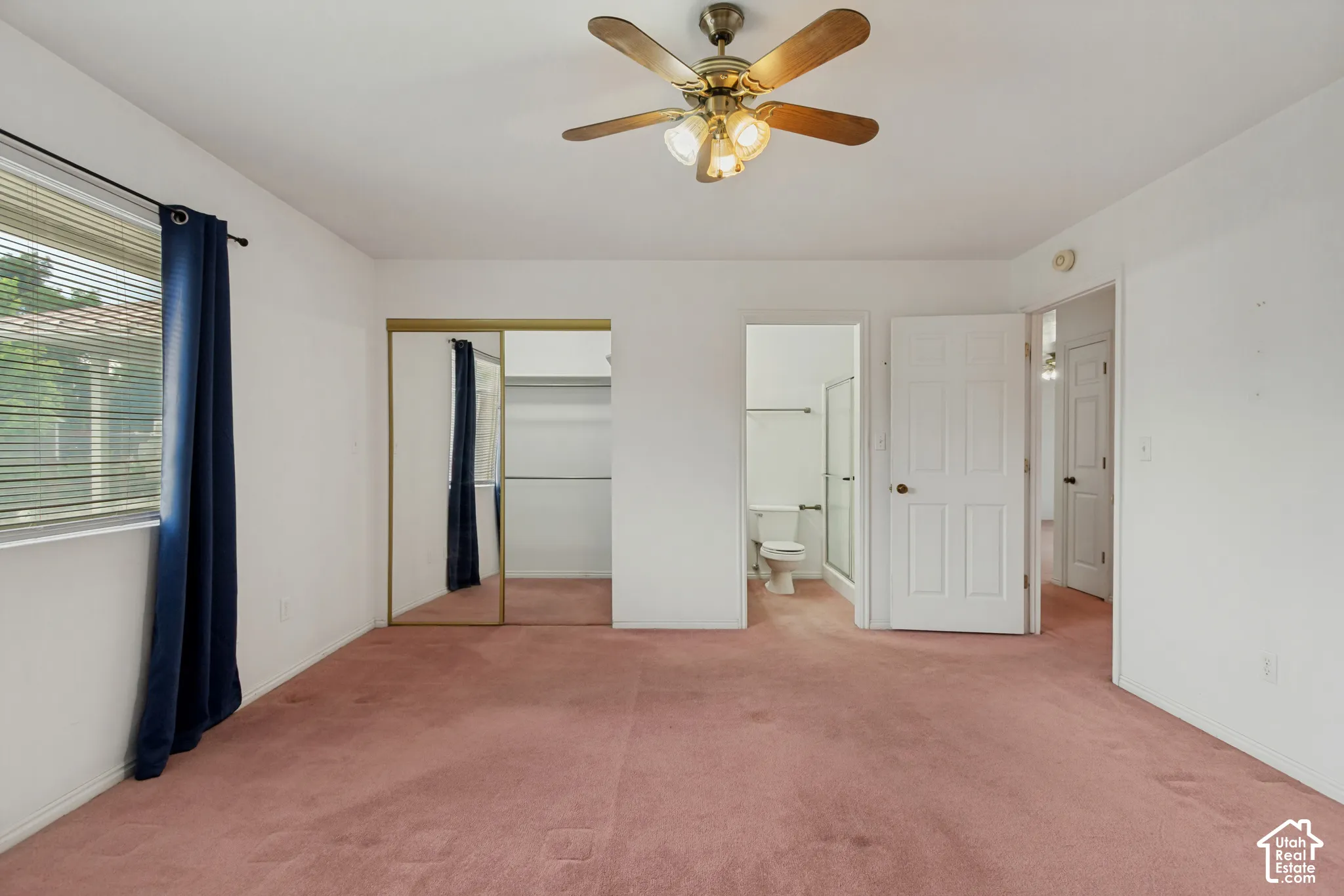 Primary bedroom shown vacant. Ceiling fan with integrated lighting, window with blinds, mirrored closet doors, and an attached bathroom with a glass-enclosed shower