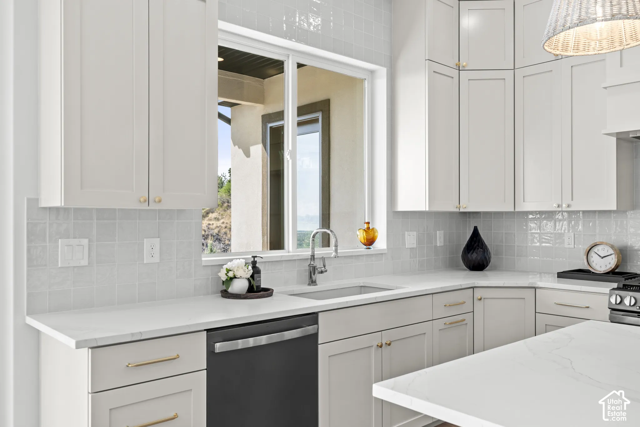 White shaker-style cabinetry with gold-tone hardware, a stainless steel sink with a gooseneck faucet, light-toned countertops, a built-in dishwasher, and a tiled backsplash