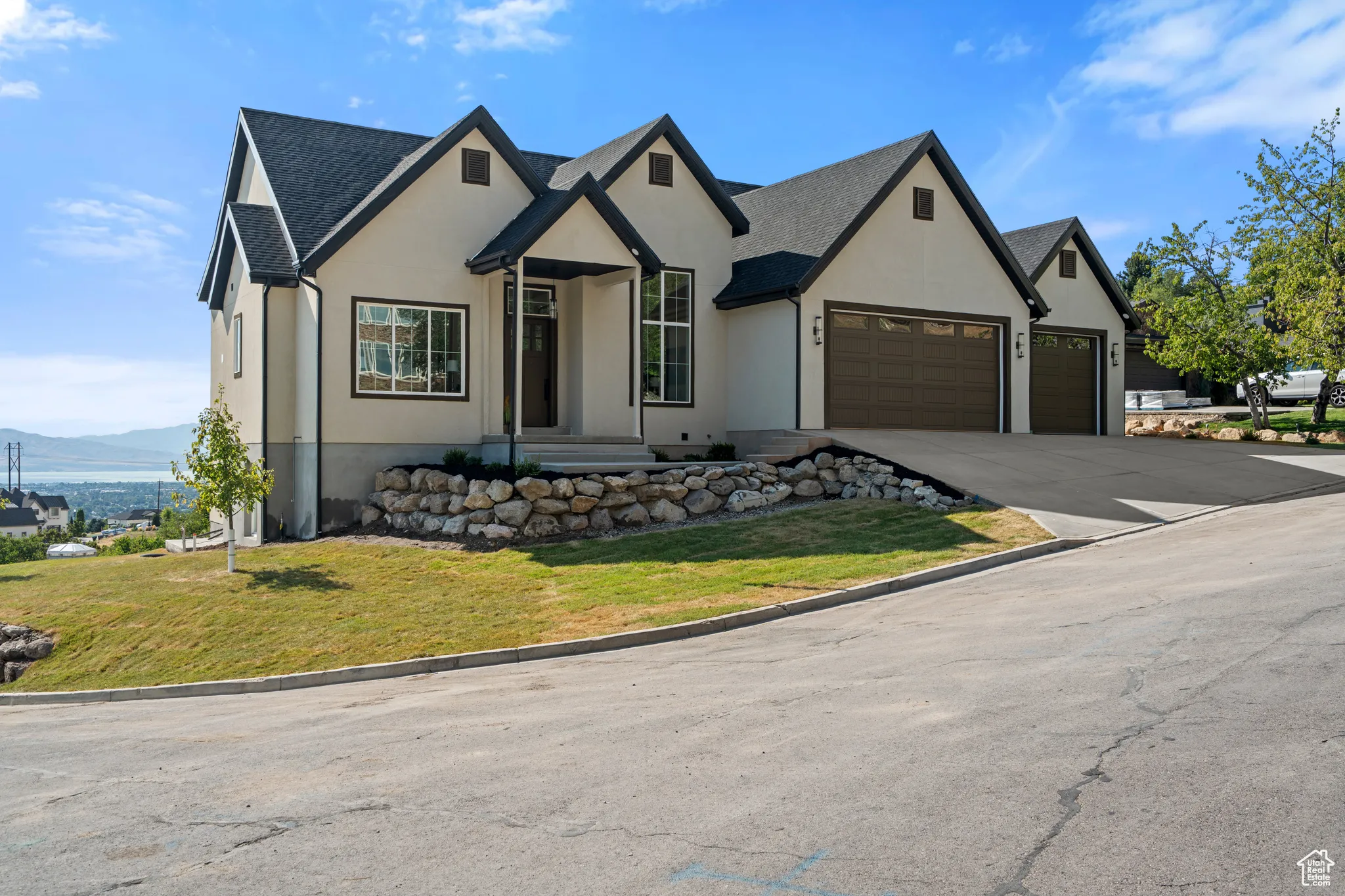Stucco exterior with dark trim accents, multi-gabled roofline, dual garage doors, concrete driveway, and a stone retaining wall