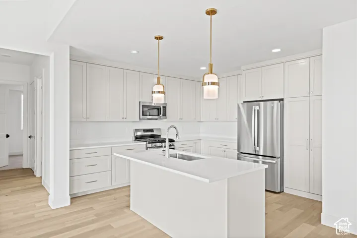 Kitchen featuring white cabinetry, stainless steel appliances, a center island with sink, pendant lighting, and wood-finish flooring