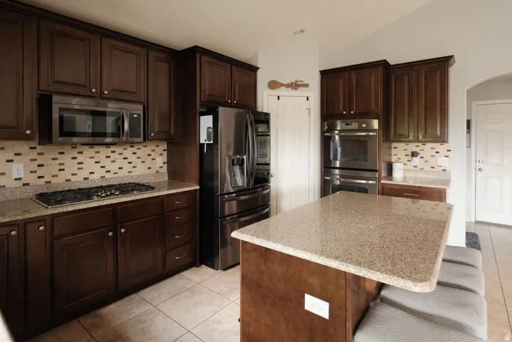 Dark wood-finish cabinetry, stainless steel appliances, patterned tile backsplash, light-toned countertop, and a kitchen island with seating
