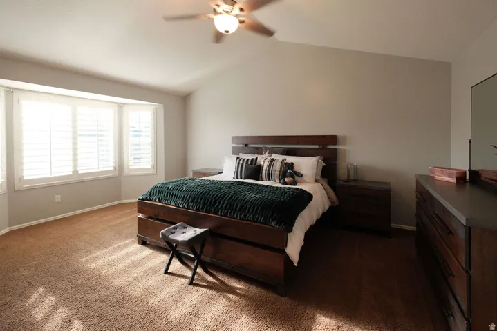 Carpeted room featuring a ceiling fan, a vaulted ceiling, and a bay window with plantation shutters