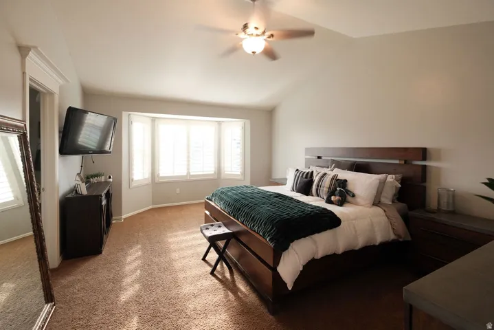 Vaulted ceiling with an attached ceiling fan, bay windows with plantation shutters, wall-mounted television, and carpet flooring