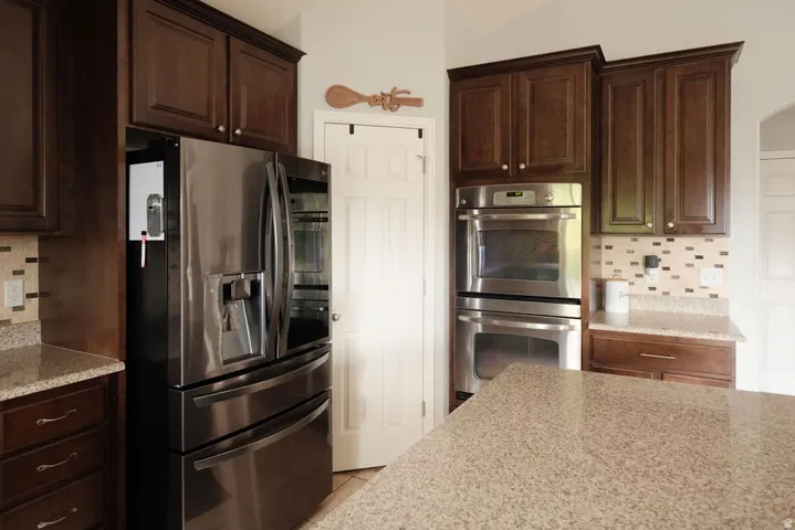 Stainless steel French door refrigerator, built-in double ovens, dark wood cabinetry, granite countertops, and mosaic tile backsplash