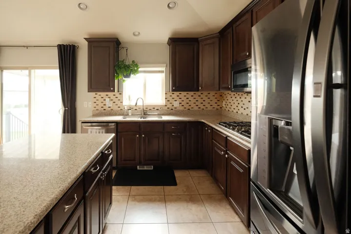 Galley kitchen featuring rich wood cabinetry, stainless steel appliances including a refrigerator and wall-mounted oven, ceramic tile flooring, light-filled windows with natural sightlines, and a mosaic tile backsplash with coordinating quartz countertops