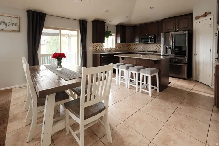 Kitchen featuring dark wood-finish cabinetry, stainless steel appliances, a center island with countertop seating, a decorative tile backsplash, and light-toned floor tiling