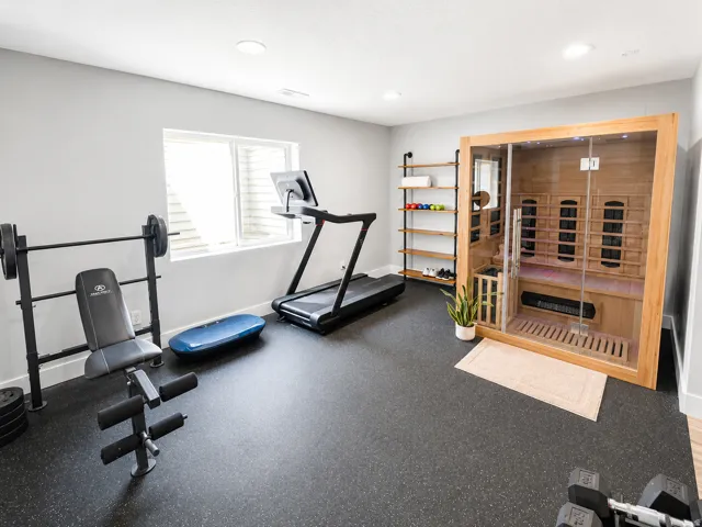 Recessed lighting illuminates a home gym with specialized black flooring, a built-in wooden sauna, and a window for natural light