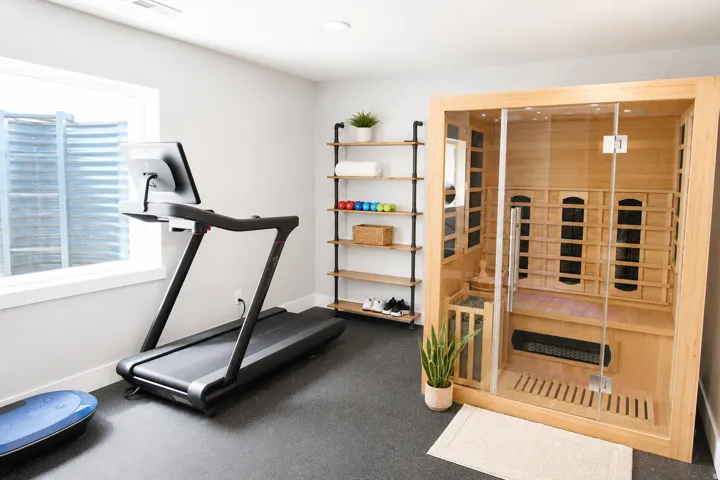 Dedicated fitness area featuring durable black rubber flooring, a built-in sauna with a glass door, and a multi-level open shelving unit