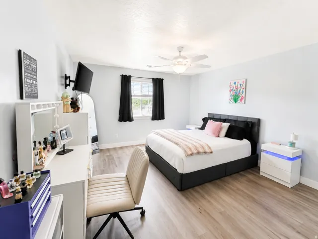 Light-toned wood-finish flooring, ceiling fan with integrated lighting, window with blinds and drapery, wall-mounted television bracket