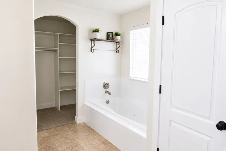 Built-in bathtub with chrome fixtures, window with blinds, tiled flooring, and an arched entryway to a closet featuring shelving and a hanging rod