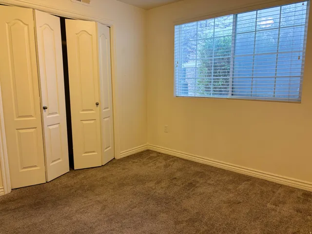 Textured carpet flooring, window with horizontal blinds, bi-fold closet doors, wall-mounted electrical outlet, and white trim molding