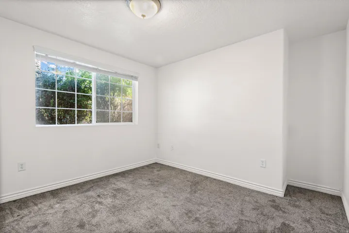 White walls with a window featuring exterior greenery, overhead flush-mount lighting, and gray carpeting