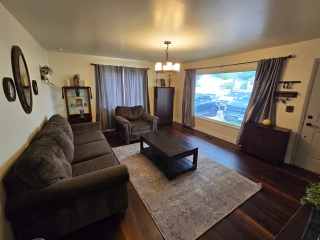 Expansive living area featuring rich wood-finish flooring, a broad picture window, and a central chandelier