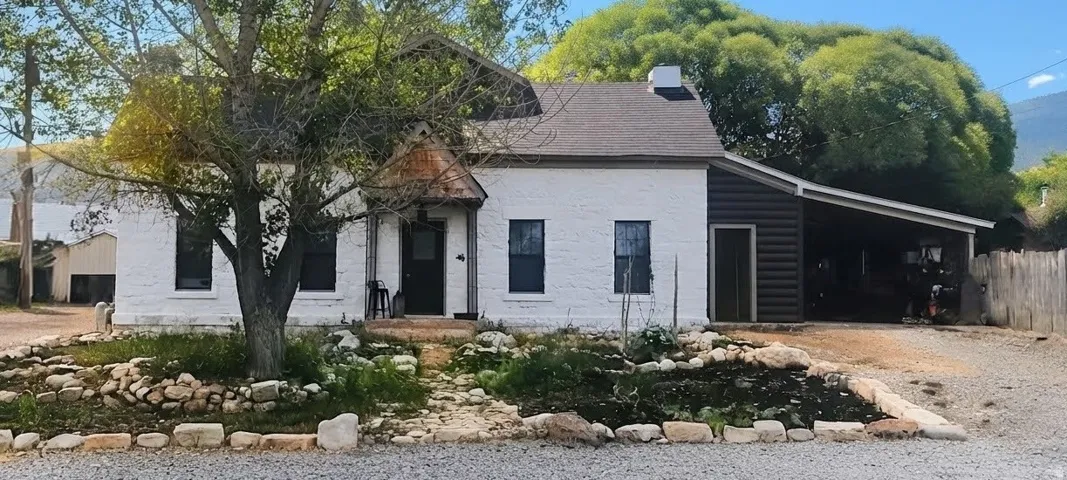 White painted brick exterior with a black shingle roof, featuring a covered carport and a stone-lined garden bed