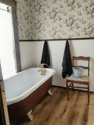 Clawfoot tub with brass fixtures, wood-finish flooring, wainscoting, and patterned wallpaper