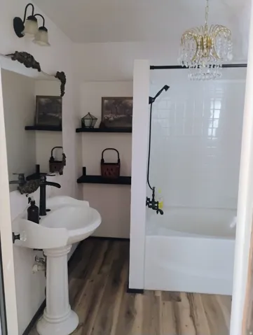 Pedestal sink with dark-toned fixtures, wood-finish flooring, built-in shelving, white subway tile shower surround, and a crystal chandelier