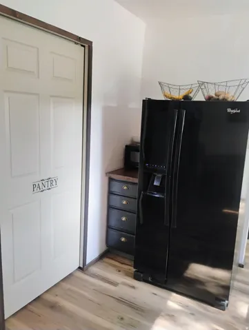 Pantry door with dark wood trim, black side-by-side refrigerator, dark wood-finish countertop, dark cabinetry with brass hardware, and wood-finish flooring