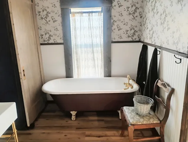Clawfoot soaking tub with brass fixtures, wood-finish flooring, half-wall wainscoting, decorative patterned wallpaper, and dark-stained window trim