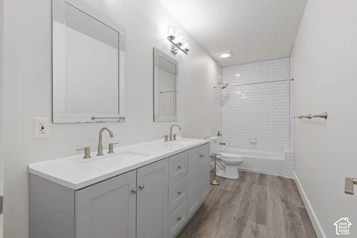 Dual vanity with a white countertop and gray cabinetry, wood-finish flooring, subway tile shower surround, and brushed nickel fixtures
