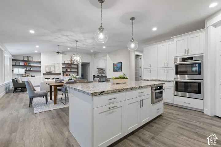 White shaker cabinetry, built-in stainless steel double ovens, central kitchen island with granite countertop, wood-finish flooring, and recessed lighting