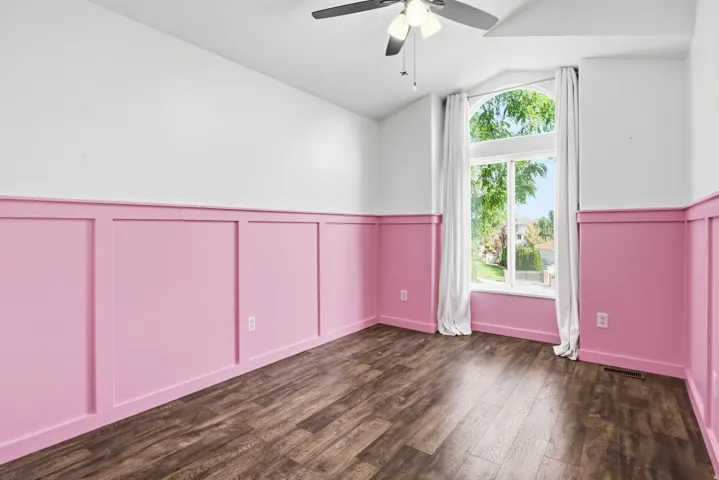 Vaulted ceiling, wood-finish flooring, accent wainscoting, large arched window, and ceiling fan with integrated lighting