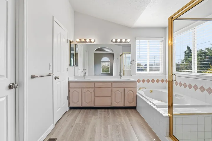 Vaulted ceiling with wood-finish flooring, dual vanity, jetted tub, and a separate shower with brass-toned framing