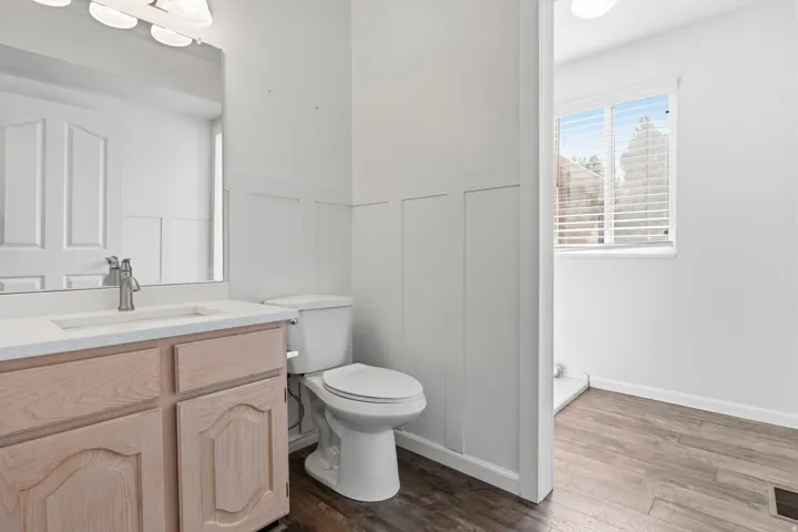 Wood-finish flooring, white board and batten wainscoting, light wood-tone vanity with a rectangular sink, wall-mounted mirror, and a window with blinds