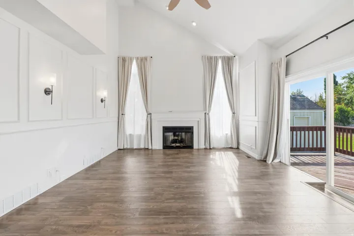 Vaulted ceiling with a ceiling fan, fireplace with a white mantel, wood-finish flooring, and sliding glass door leading to a wood deck